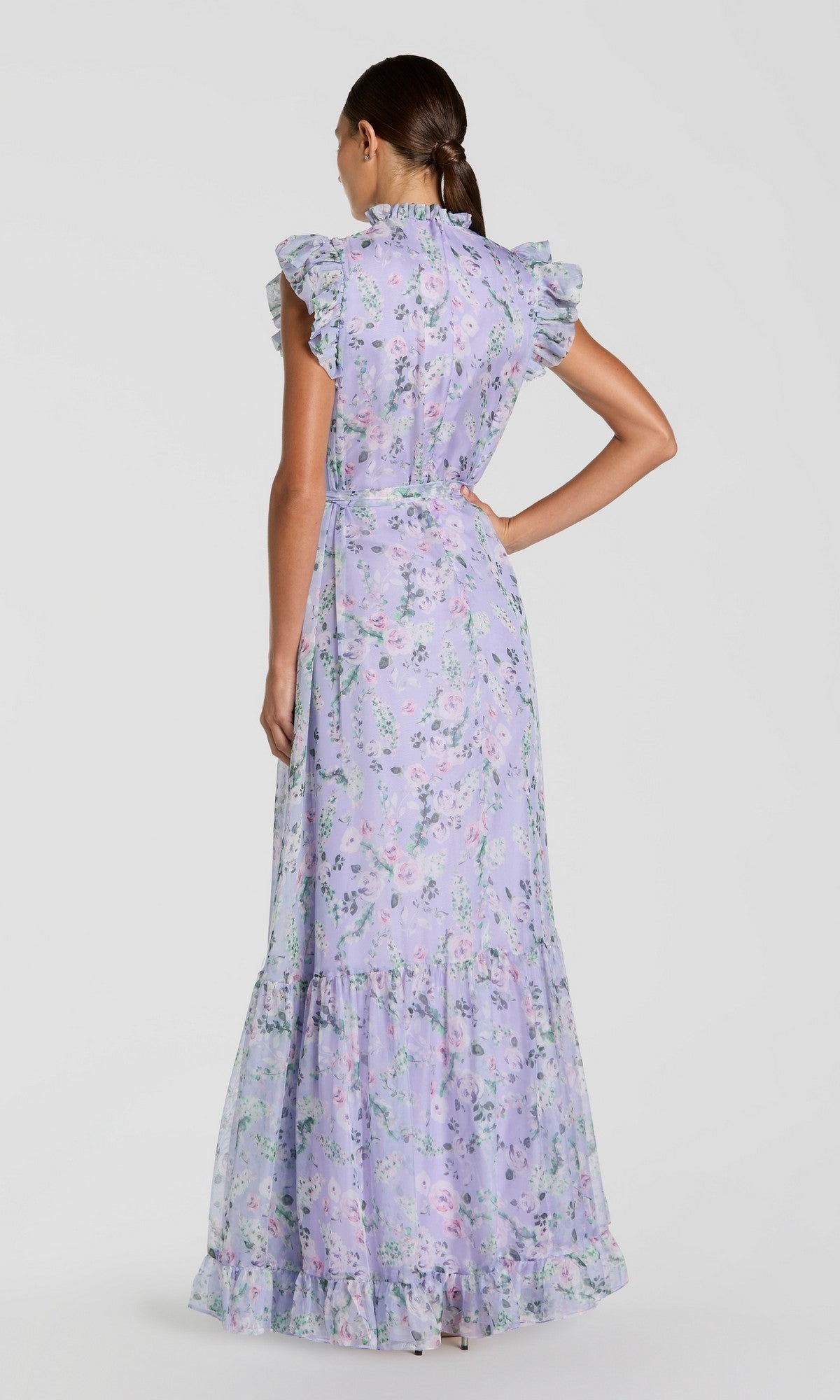 A woman with a low ponytail is seen from the back wearing the Mac Duggal 56116 Lilac Floral-Print Formal Gown, a sleeveless long dress with ruffle details, set against a plain background.